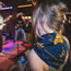 A woman with blonde hair and a blue flowered scarf from the Howler Bandanas Yellow Rose Collection watches a live band perform on stage at the ACL Festival, holding a can drink as colorful lights illuminate the musicians.