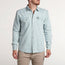 A man wearing a rugged denim shirt with two chest pockets and beige pants stands against a plain, light-colored background, showcasing a versatile style as he turns his head slightly to the side.