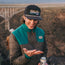 A smiling person in a green and brown jacket and a black Hustler soft crown cap with an adjustable snapback closure stands outdoors holding a Modelo can, with a canyon bridge in the background.