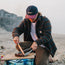 A person wearing an unstructured snapback with a soft crown and a striped jacket slices cooked meat on a wooden board at a sandy beach, with houses and the ocean in the background.