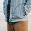 A person stands on the beach with hands in the pockets of a light blue Depot Jacket featuring a subtle lightning bolt stitch, wearing a green patterned shirt underneath and tan pants. The ocean and sand are visible in the blurred background.