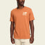 A man wearing a burnt orange Howler blended t-shirt with a small white Howler Brothers logo on the left chest, standing against a plain light background. The cotton-poly blend ensures maximum comfort, paired with light-colored pants.