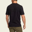 A man is standing with his back to the camera, wearing a black vintage t-shirt and olive green pants against a plain, light background.
