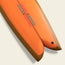 Close-up of the tail of an orange Almond Surfboards surfboard with a split fish tail design, brown edges, and blue and red text reading Lightning Powered Surf Machine printed on the board. The surfboard is set against a plain light background.