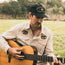 A man wearing a beige shirt and a black unstructured snapback with a soft crown sits outdoors on grass, playing an acoustic guitar. Trees and bushes are in the background.