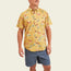 A man wearing the Mansfield Shirt, a yellow, short-sleeve, button-up Hawaiian shirt with unique seasonal patterns and blue shorts stands smiling with his hands relaxed at his sides against a plain light background.