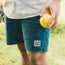 A person wearing a light patterned shirt and teal Pressure Drop Cord Shorts made from stretch cotton corduroy stands outside on grass, holding two yellow citrus fruits. The comfortable shorts feature a patch with a palm tree design on the left leg.