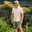 A man wearing a light patterned short-sleeve shirt, Quonset Camp Shorts with vintage-style buttons, and a gray cap stands smiling among green plants in front of a dark building with large windows.