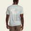A person wearing a light blue Howler blended t-shirt featuring a pelican graphic and text, Heed the Call, Est. 2010, Austin Texas, USA, viewed from the back. Made from a soft cotton-poly blend for maximum comfort.