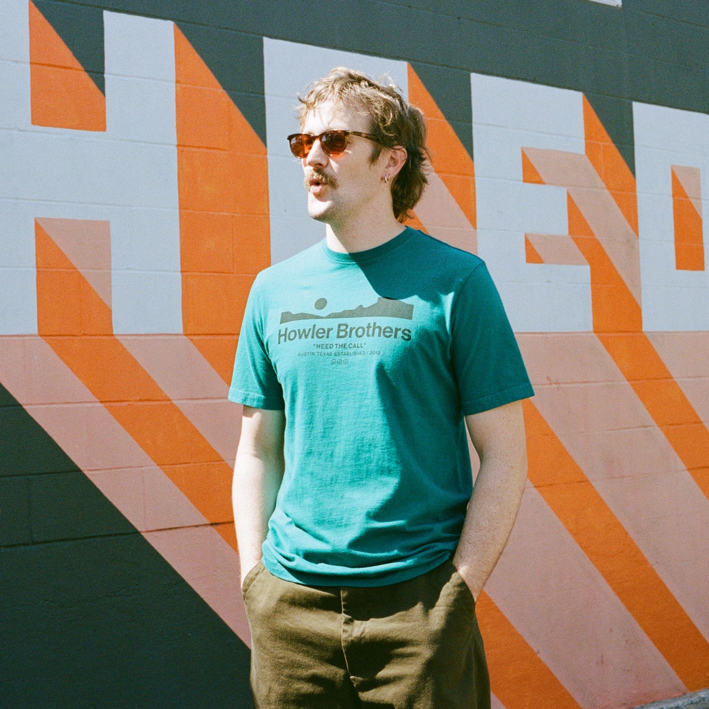 A man with light brown hair, sunglasses, and a teal Howler Brothers T-shirt stands against a colorful mural featuring bold geometric shapes and large orange and white letters, echoing the vibe of experimental artwork. His hands are in his pockets. - View 2