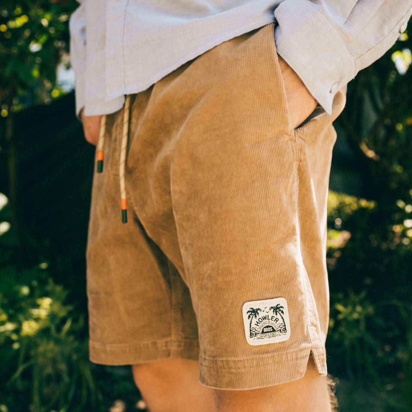 A person wearing tan Pressure Drop Cord Shorts made from stretch cotton corduroy with a palm tree patch on the left leg stands outdoors, hands in pockets, enjoying all day comfort. Greenery and sunlight fill the background. - View 2