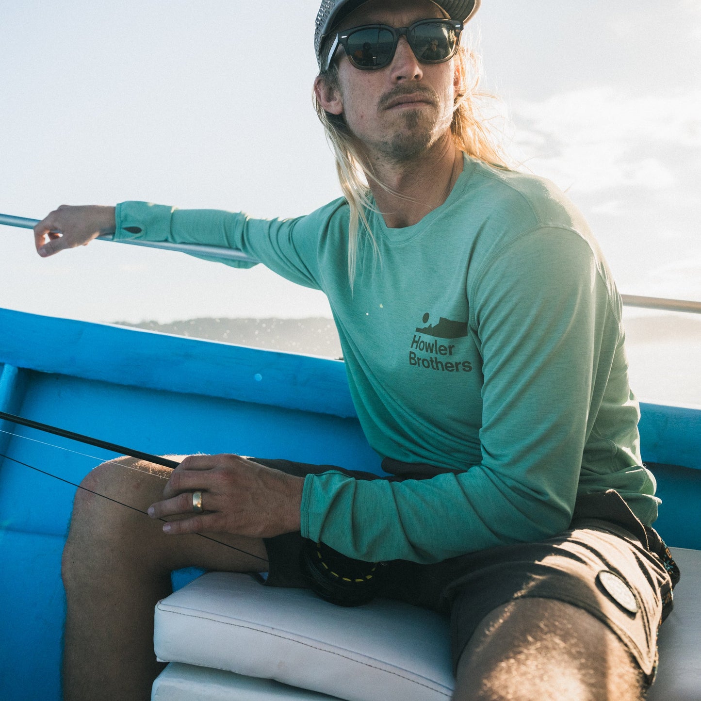 A man wearing sunglasses, a cap, and a green Howler Brothers UPF 50+ shirt sits on a blue boat holding fishing equipment, looking off to the side in bright sunlight—ready for any outdoor activities. - View 2