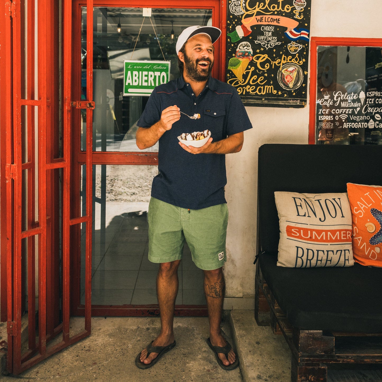A man in Pressure Drop Cord Shorts, a polo shirt, and a cap stands by a red door, smiling and eating ice cream. Behind him are colorful signs and pillows on a bench that say “Enjoy Summer Breeze.”. - View 2