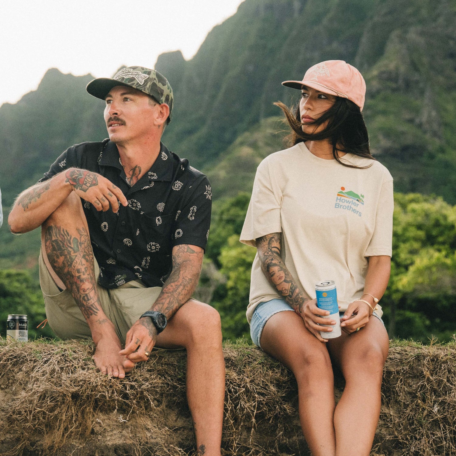 A man and woman with tattoos sit outdoors on a grassy ledge, sporting Howler blended t-shirts for maximum comfort. The woman holds a canned drink, while lush green mountains provide a stunning backdrop. - View 6