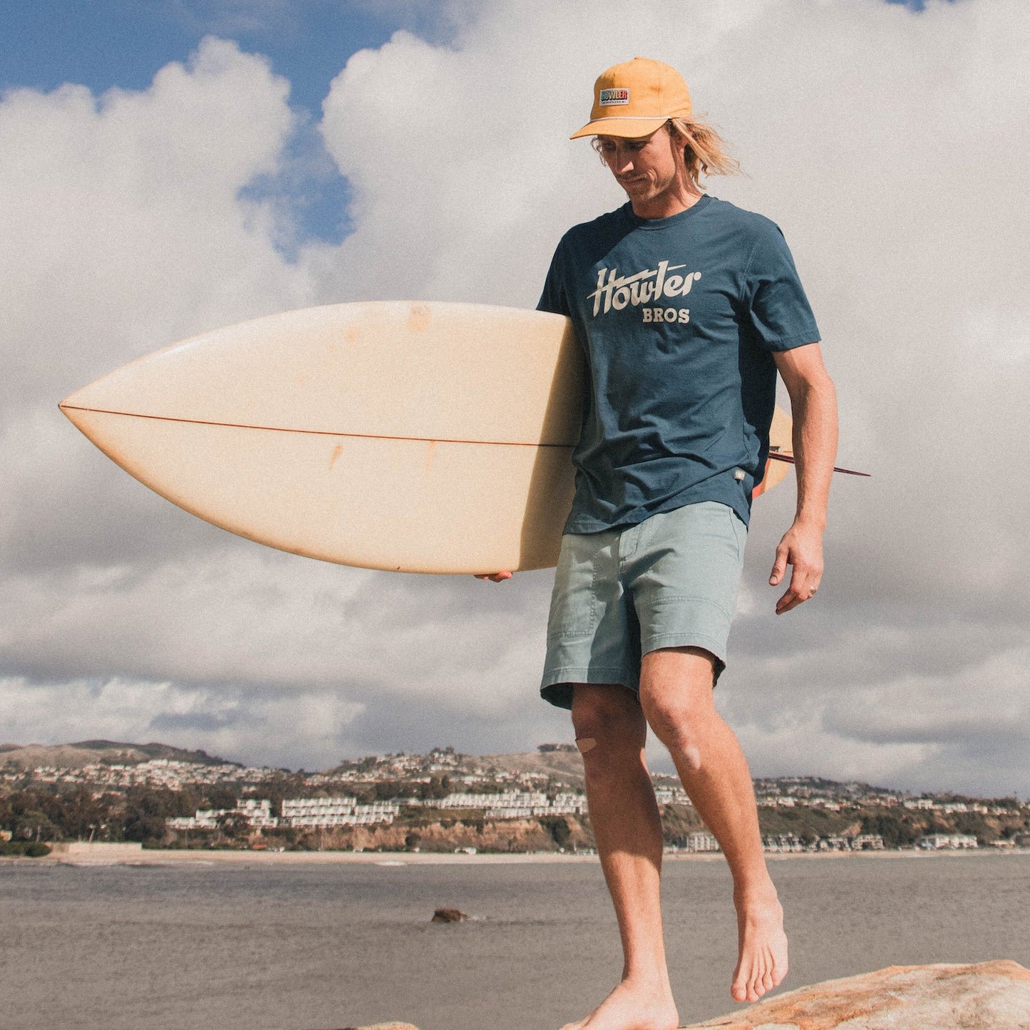 A man wearing a yellow cap, classic Howler designs blue cotton-poly blend t-shirt, and light green shorts walks barefoot on rocks by the water, carrying a surfboard with a coastal town and cloudy sky in the background. - View 2