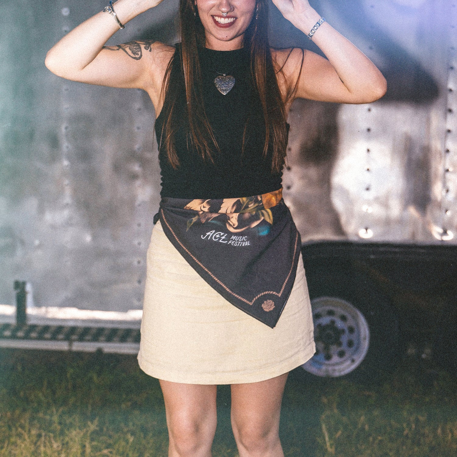 A woman in a black sleeveless top and beige skirt stands outdoors, smiling, with a Howler Bandanas scarf reading ACL Music Festival tied around her waist. She has long hair, bracelets, and a heart-shaped necklace. - View 4