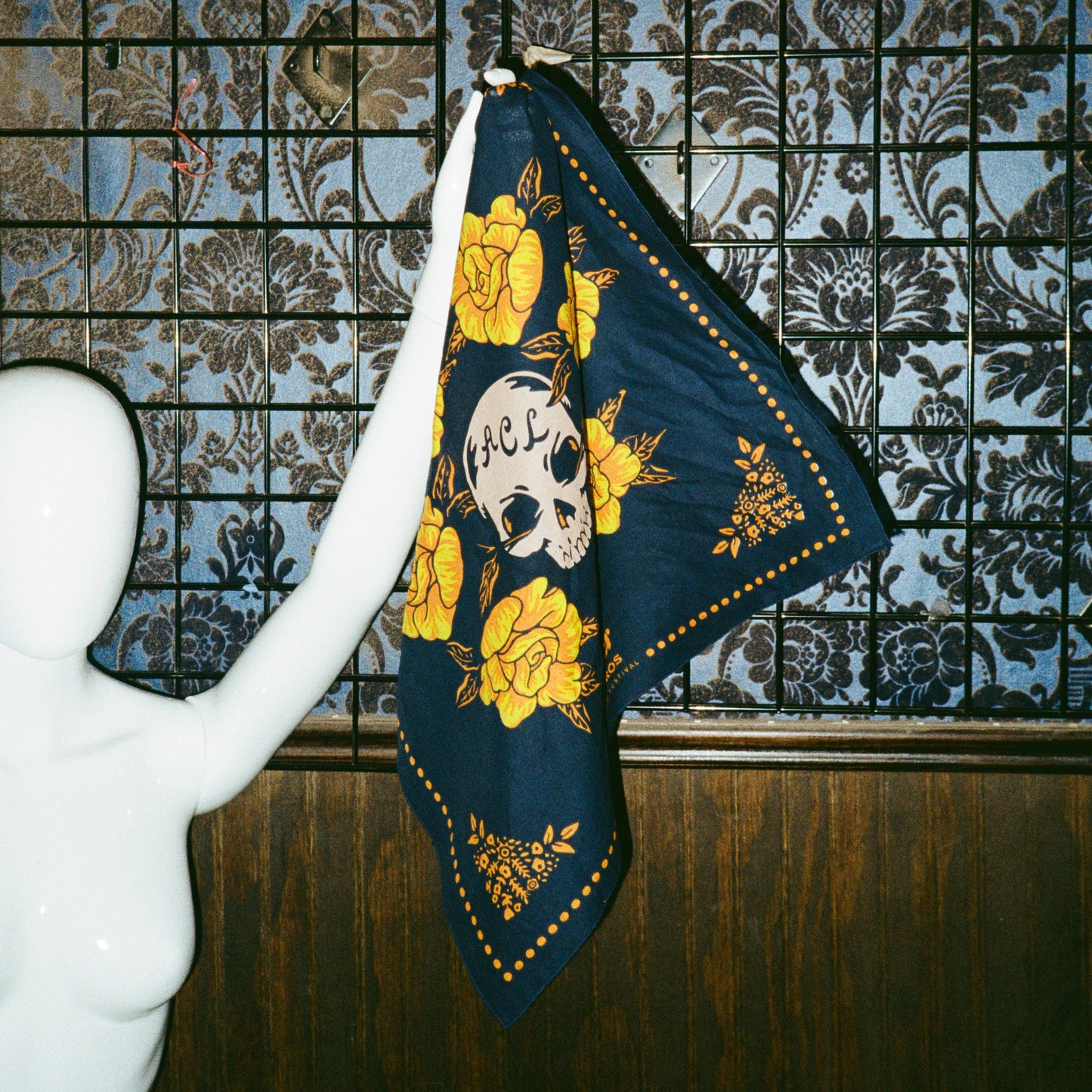 A white mannequin holds up a navy blue scarf from the Yellow Rose Collection, decorated with yellow flowers and a skull with RIP on its forehead. The background features patterned wallpaper and a wooden panel. - View 7