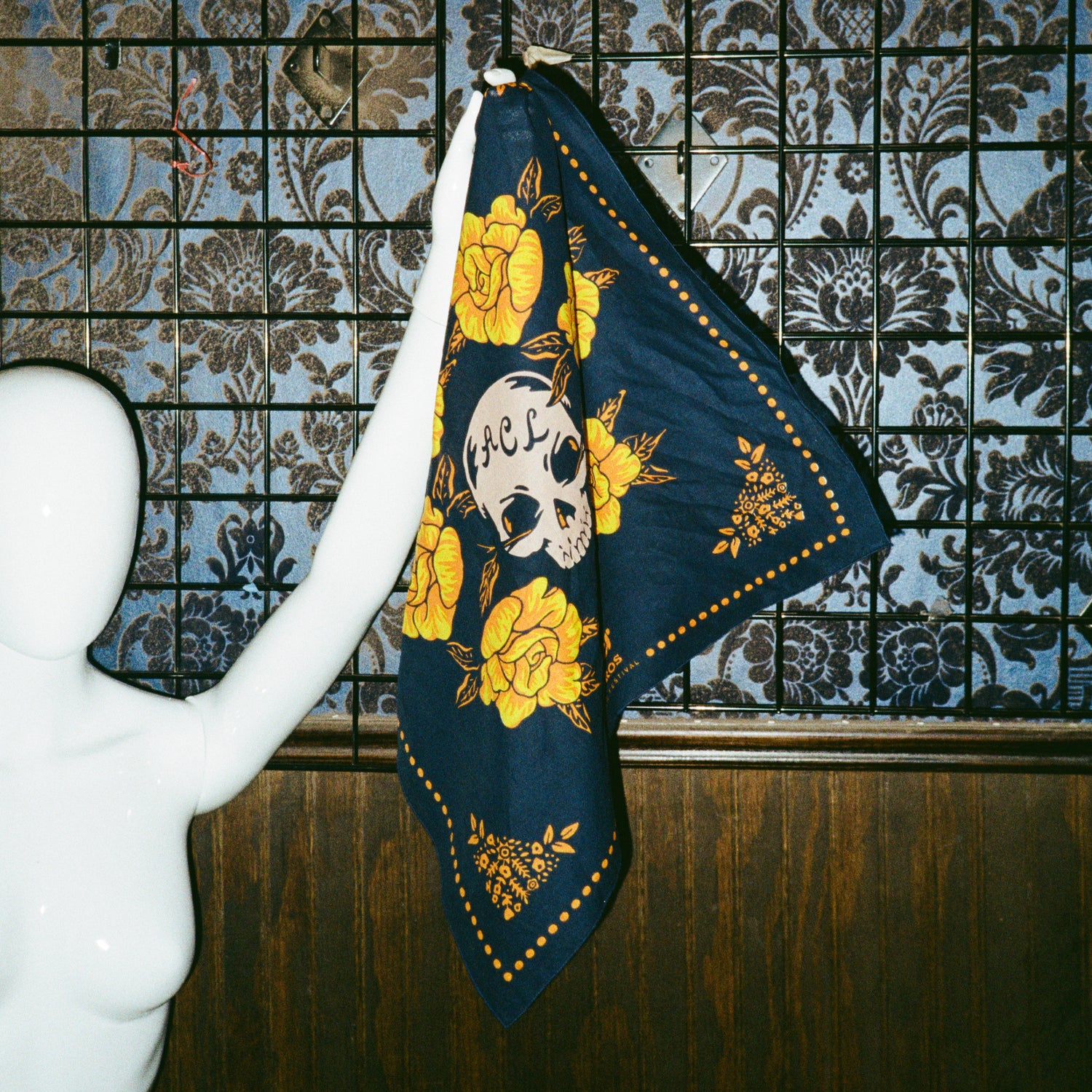 A white mannequin holds up a navy blue scarf from the Yellow Rose Collection, decorated with yellow flowers and a skull with RIP on its forehead. The background features patterned wallpaper and a wooden panel. - View 7