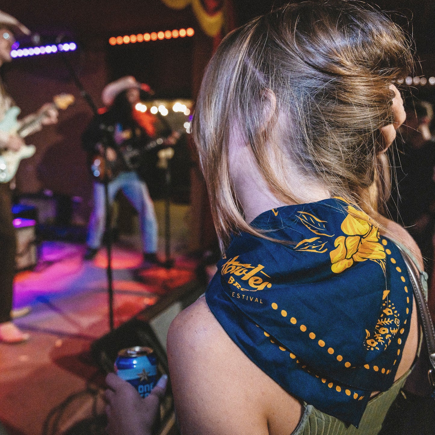 A woman with blonde hair and a blue flowered scarf from the Howler Bandanas Yellow Rose Collection watches a live band perform on stage at the ACL Festival, holding a can drink as colorful lights illuminate the musicians. - View 6