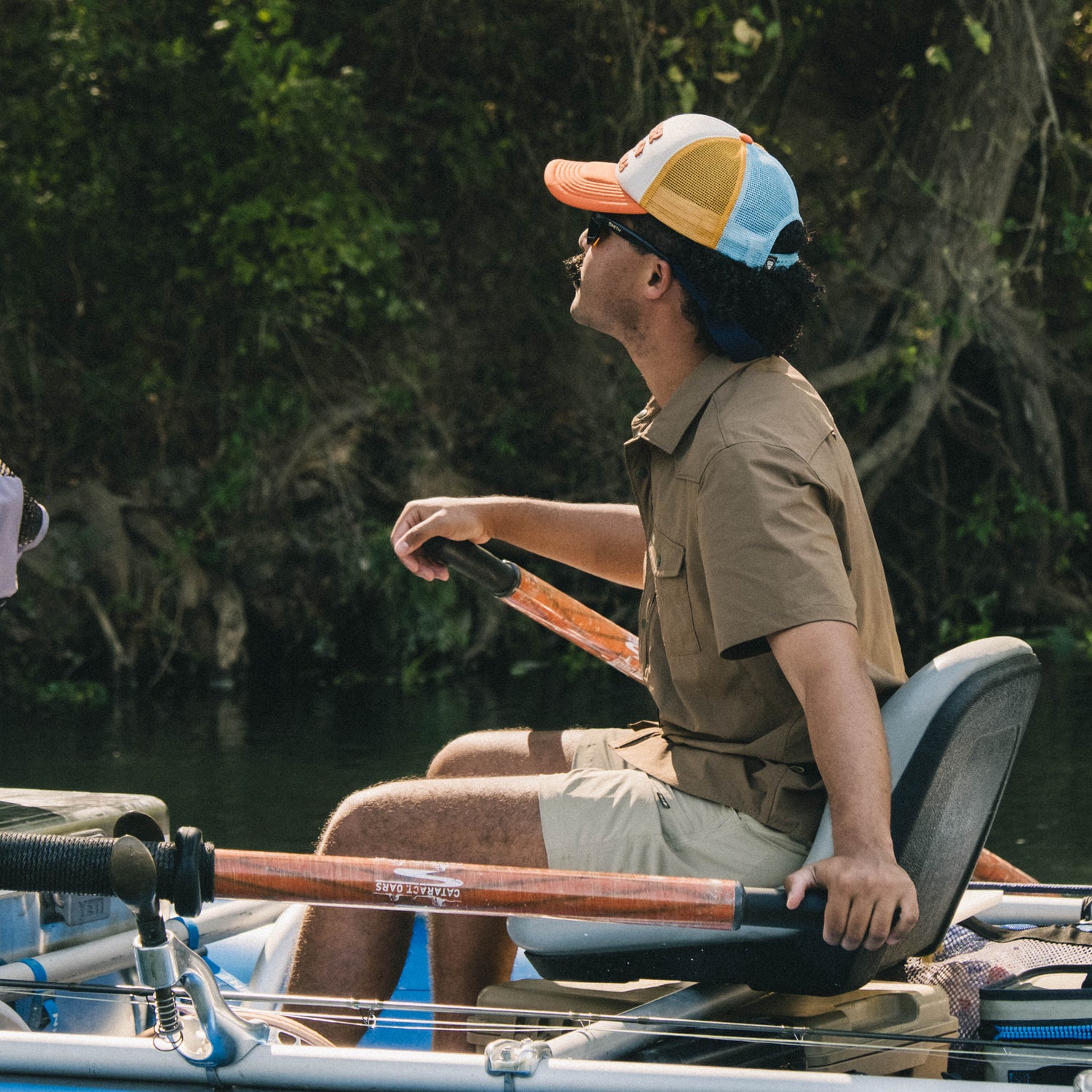 A person wearing a vintage-inspired hat and sunglasses sits in a boat, holding oars, rowing on a calm body of water surrounded by green foliage. - View 3