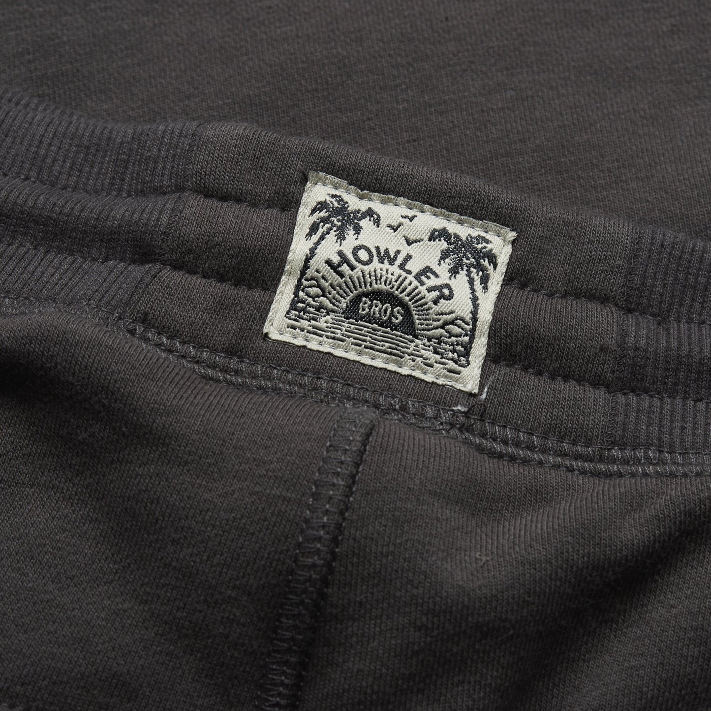 Close-up of a black shirt with a white fabric label sewn inside the collar, featuring the Howler Bros logo with palm trees, waves, and sun rays—perfect to pair with Mellow Mono Sweatpants or classic French terry sweatpants. - View 6