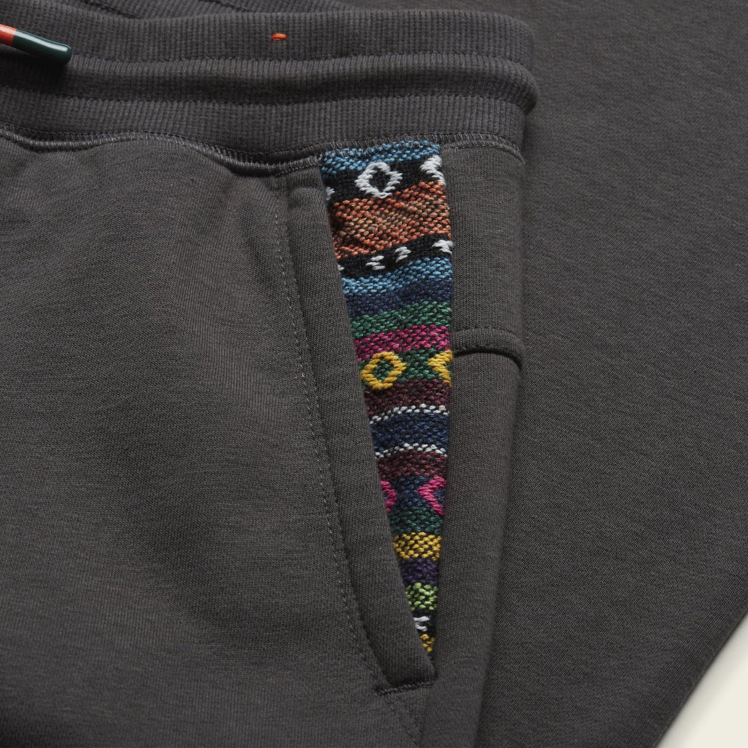 Close-up of dark gray Mellow Mono Sweatpants featuring a pocket with colorful, geometric patterned trim in blue, orange, green, pink, yellow, and white and a comfortable drawstring waistband. - View 4