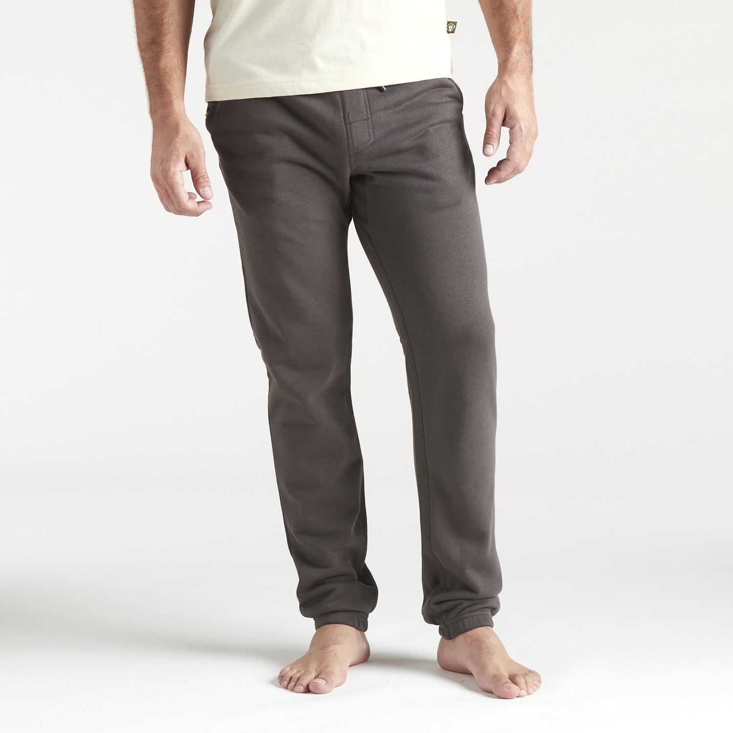 A person stands barefoot, wearing fitted dark gray French terry sweatpants with a drawstring waistband and a light-colored shirt, arms relaxed by their sides against a plain white background. The photo is cropped at the shoulders. - View 2