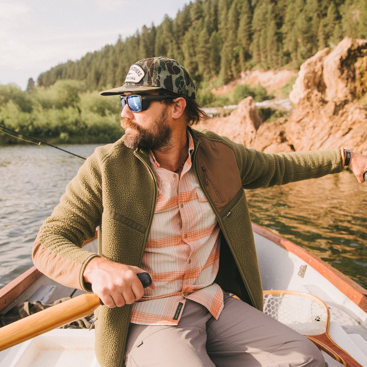A man wearing sunglasses, a cap, and a fleece jacket rows a small boat on a lake surrounded by trees and rocky hills, looking to the side. - View 2