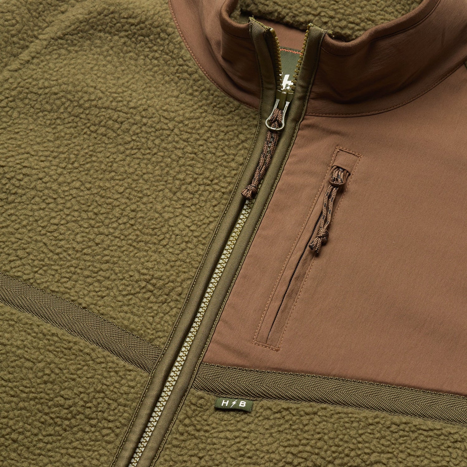 Close-up of an olive green and brown polyester fleece jacket with a zipper, a chest pocket featuring a zipper pull, and a small H/B tag near the main zipper. The soft, textured fabric makes it perfect for fall outerwear. - View 3