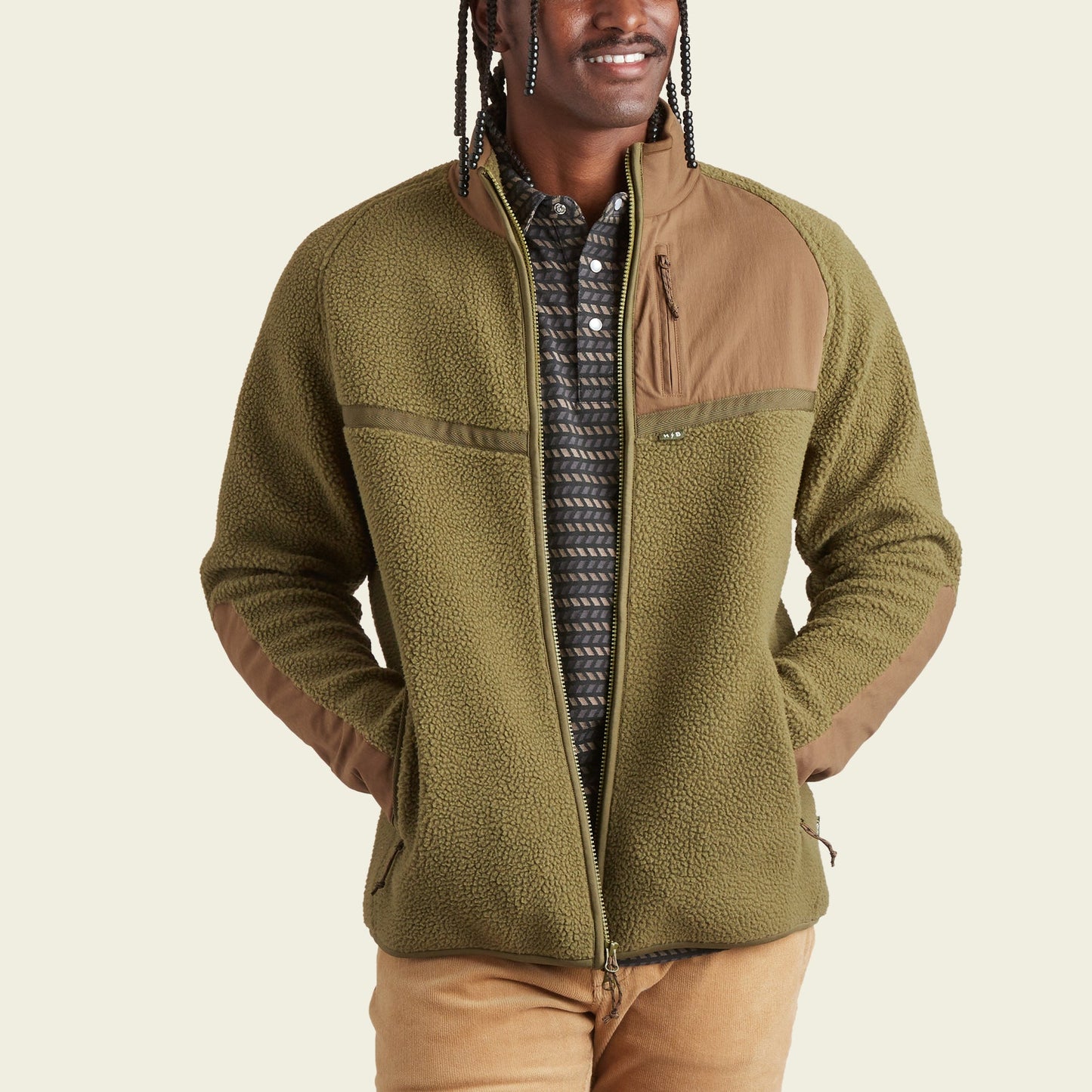 A person wearing an olive green fleece jacket with tan fabric accents on the shoulders and pockets, a patterned shirt underneath, and tan pants stands with hands in jacket pockets, smiling—ready for stylish fall outerwear. - View 4