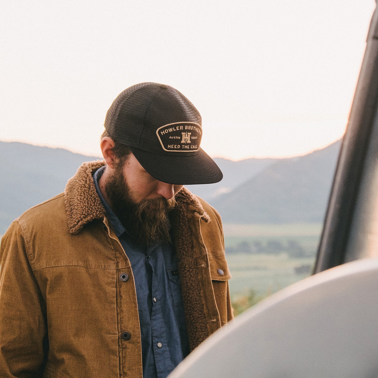 A bearded man in a black cap and corduroy jacket with sherpa lining looks down, standing outdoors with mountains and a soft, pastel-colored sky in the background. - View 9