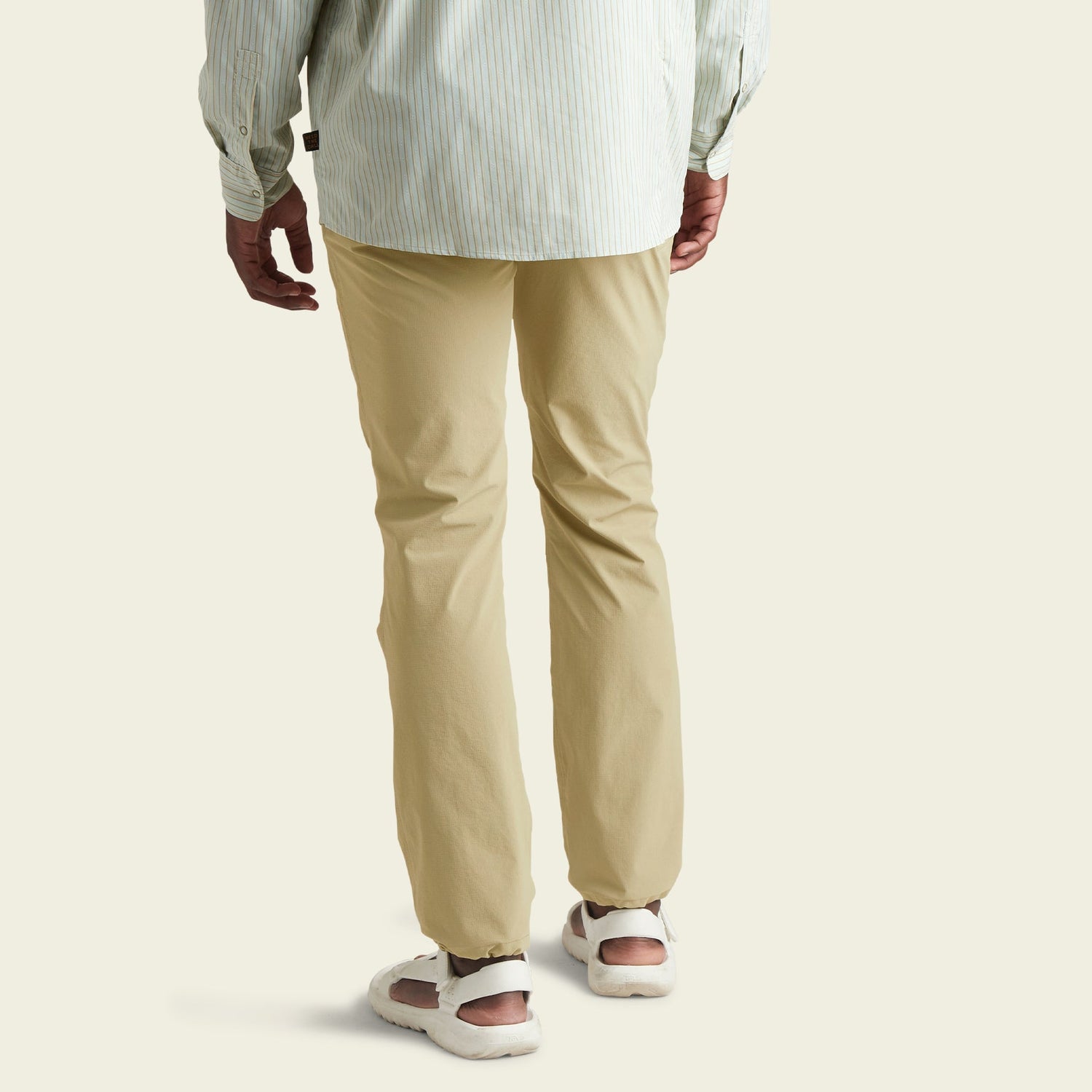 A person wearing light beige tech pants, a light striped long-sleeve shirt, and white sneakers, shown from the back, standing against a plain light background. - View 4
