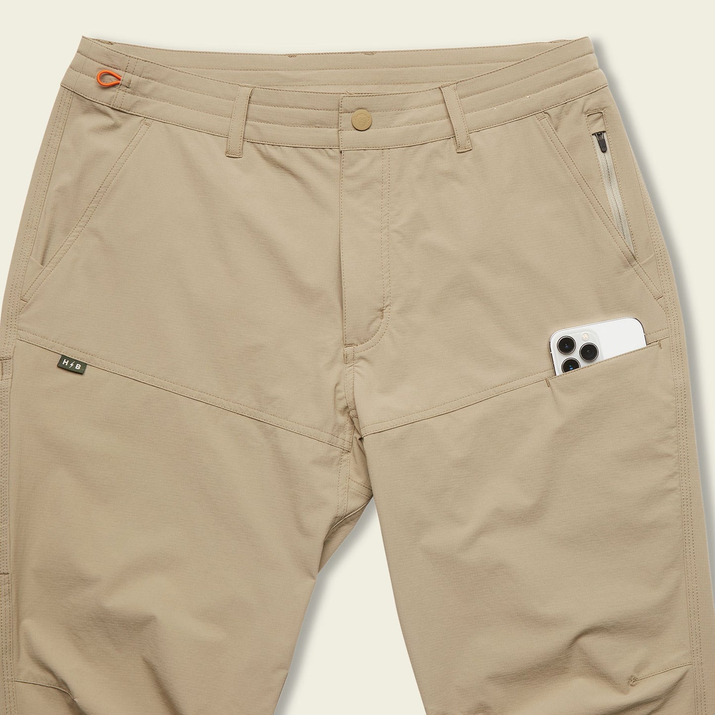 Beige tech pants with multiple pockets, including a zippered pocket and a large side pocket holding a white smartphone. These stretch nylon pants feature a button closure and belt loops for comfort and functionality. - View 9