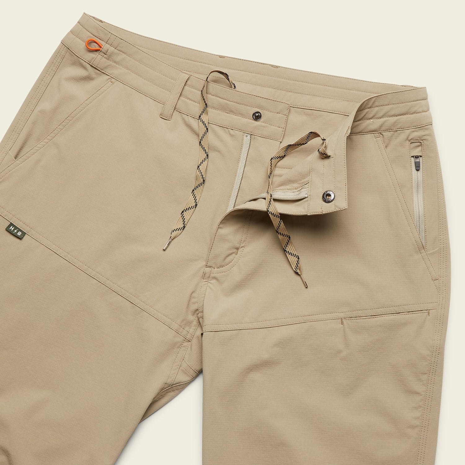 Beige stretch nylon Tech pants with a partially open waistband showing a button, zipper, and drawstring closure. The pants feature front zippered pockets and visible stitching details. - View 5