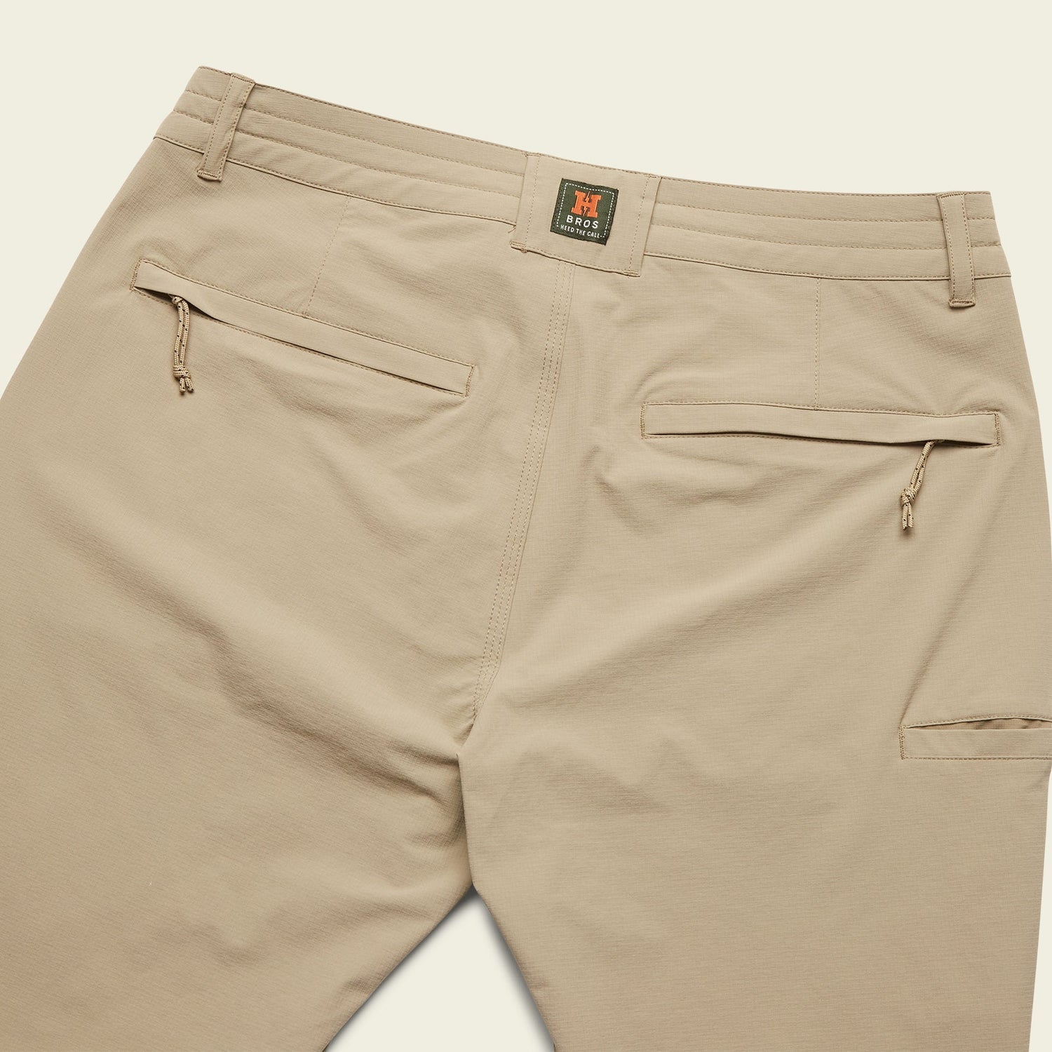 Khaki Tech pants shown from the back, featuring two zippered back pockets, belt loops, and a small stitched logo patch above the right pocket. - View 7