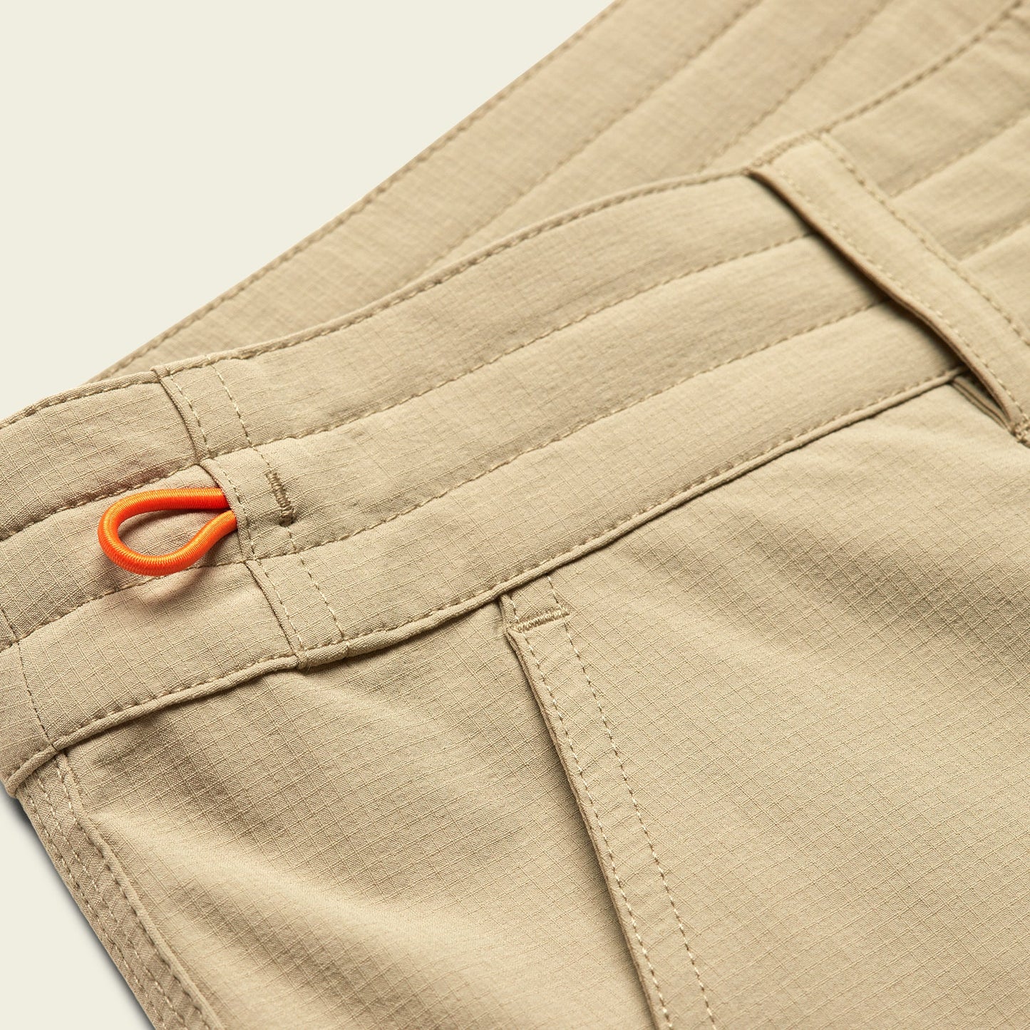 Close-up of tan Tech pants with visible stitching, belt loops, side pocket, and an orange fabric loop detail near the waistband. - View 8