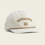 A cream-colored unstructured snapback cap with HERMANOS embroidered in yellow and brown above a stylized H logo. The soft crown and adjustable snapback closure ensure a comfortable fit, while the cap features a brown trim along the brim.
