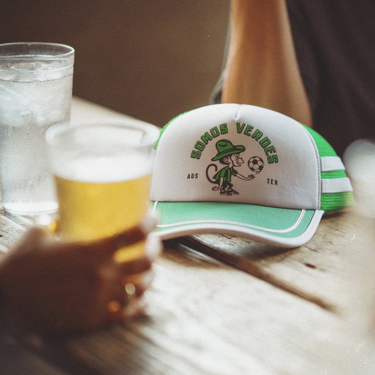 A vintage-inspired hat from the Verde Collection, this green and white trucker displays a cartoon character and SIMOS VERDES AUS TEX, resting beside a glass of water and an Austin FC fan holding a beer. - View 4