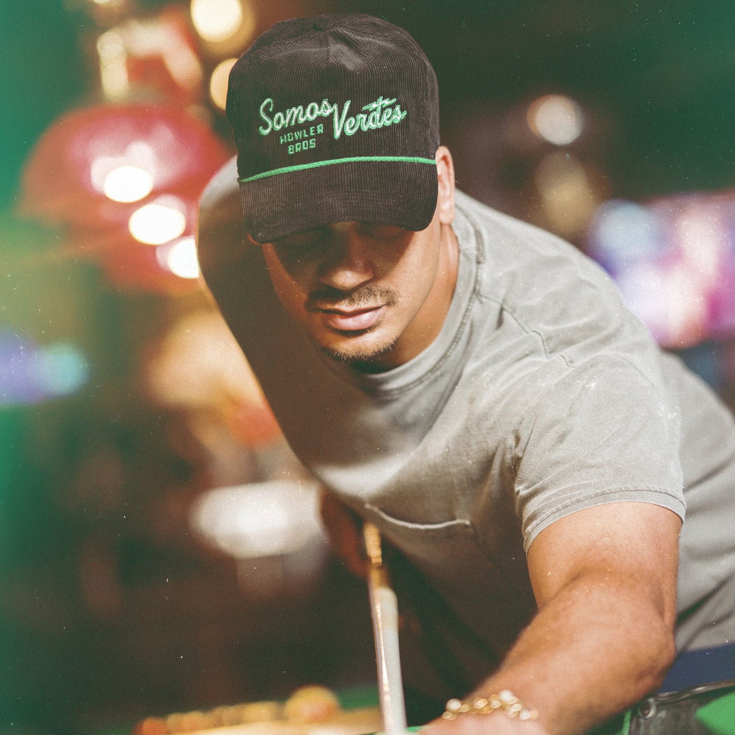 A man wearing a black Austin FC Unstructured Snapback that reads Somos Verdes and a grey t-shirt leans forward, aiming a pool cue in a dimly lit, colorful pool hall. - View 2