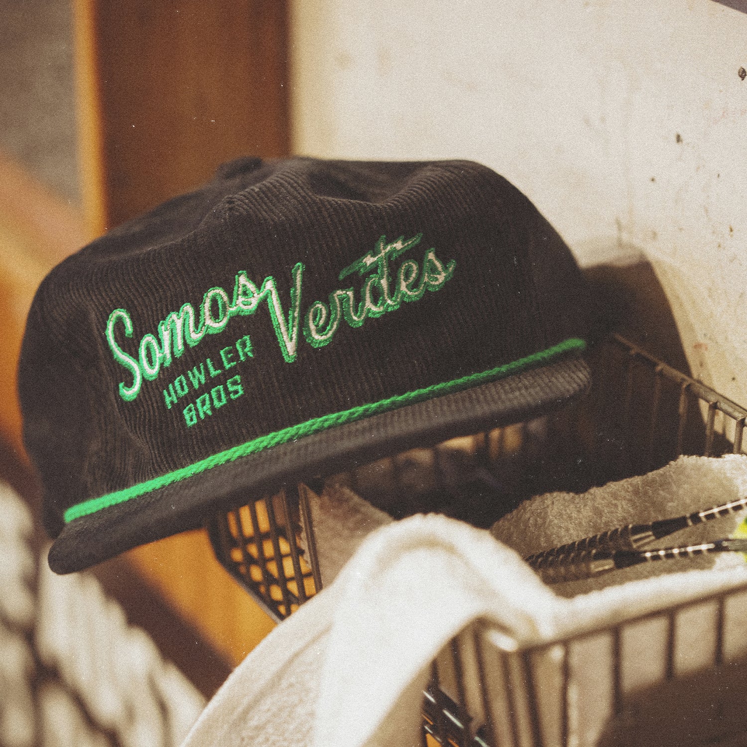 A black corduroy cap from the 2024 Verde Collection with green embroidery reading Somos Verdes HOWLER BROS rests on top of a wire basket filled with towels. - View 4