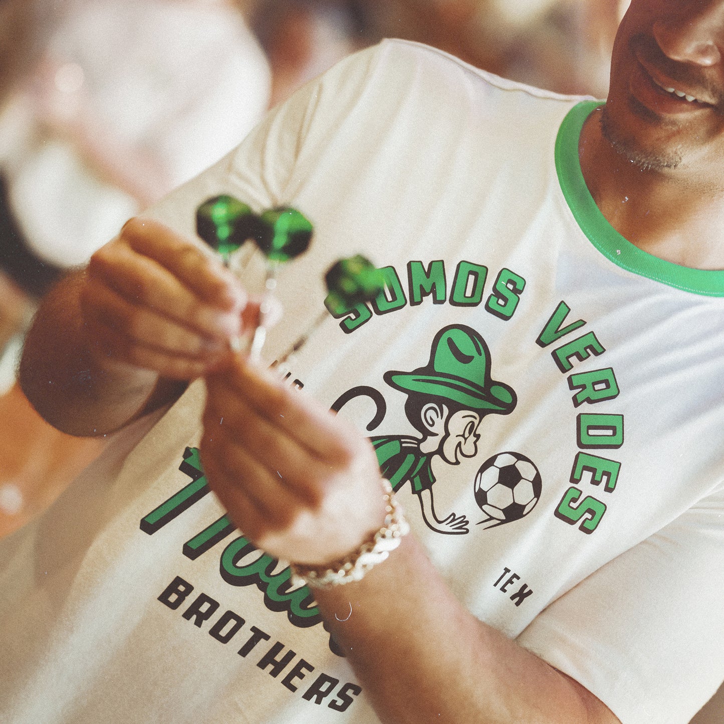 A smiling person wears a vintage t-shirt from the Verde Collection, featuring a soccer-themed design and SOMOS VERDES. They hold two green wrapped candies, showing support for Austin FC with the cartoon figure and soccer ball on their shirt. - View 2