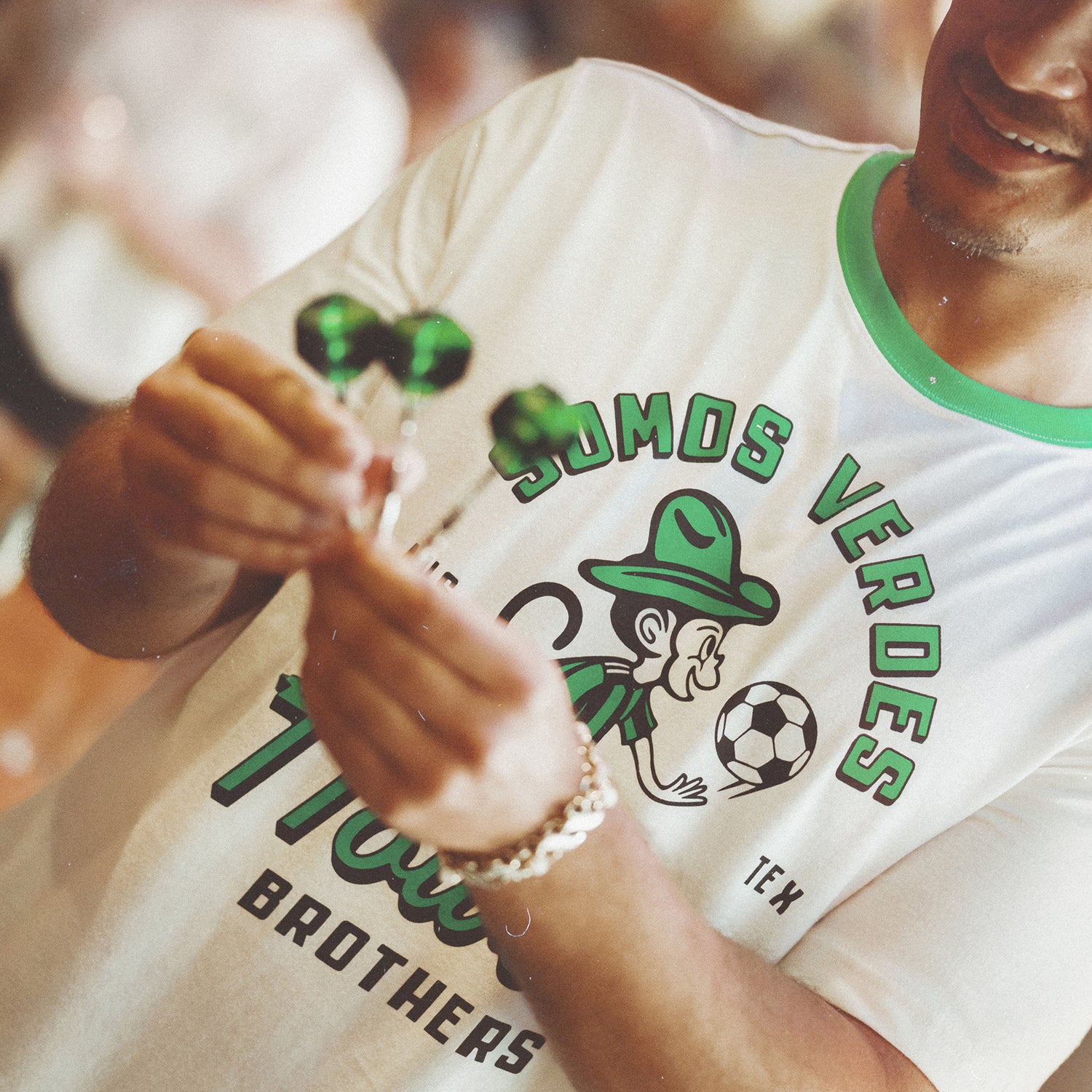 A smiling person wears a vintage t-shirt from the Verde Collection, featuring a soccer-themed design and SOMOS VERDES. They hold two green wrapped candies, showing support for Austin FC with the cartoon figure and soccer ball on their shirt. - View 2
