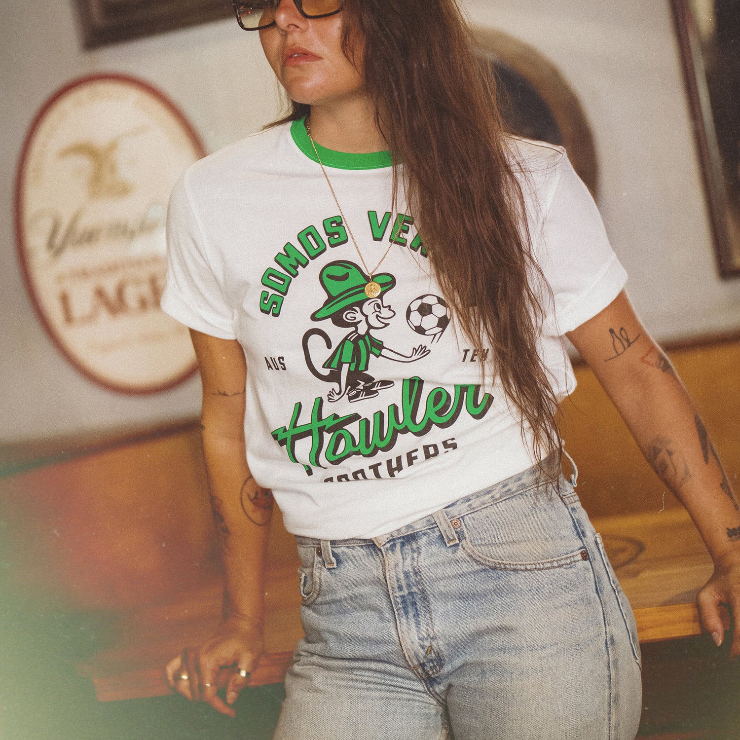 A woman in glasses and a white Austin FC vintage t-shirt from the Verde Collection, featuring a cartoon monkey, soccer ball, and the text “SOMOS VERDES” and “Howler Brothers,” stands indoors with one hand on her waist and long brown hair over her shoulder. - View 4