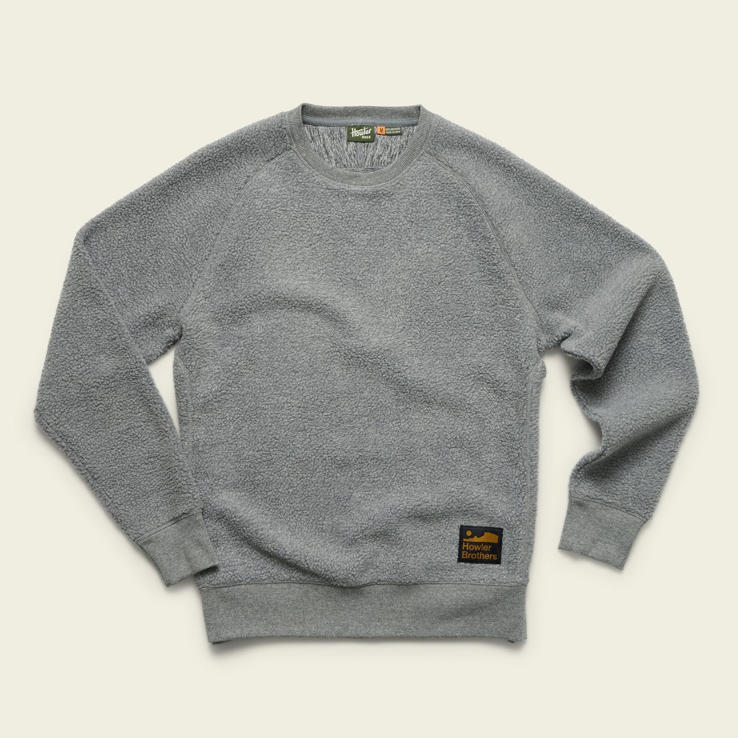 A light gray, long-sleeve crewneck sweatshirt with a soft, textured fabric. It features ribbed cuffs and hem, and a black and gold “Howler Brothers” patch on the lower left front. The shirt is laid flat on a cream background. - View 4