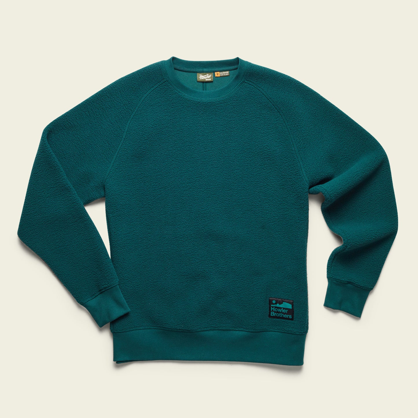 A teal fleece crewneck sweatshirt with ribbed cuffs and hem, set against a light beige background. A black and blue rectangular Howler Brothers patch is visible on the lower left side near the hem. - View 6