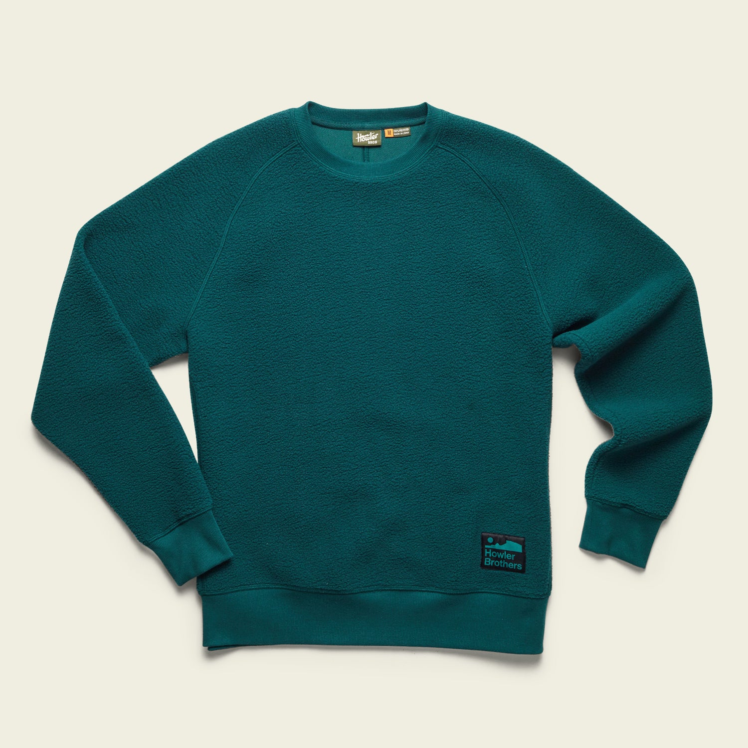 A teal fleece crewneck sweatshirt with ribbed cuffs and hem, set against a light beige background. A black and blue rectangular Howler Brothers patch is visible on the lower left side near the hem. - View 6