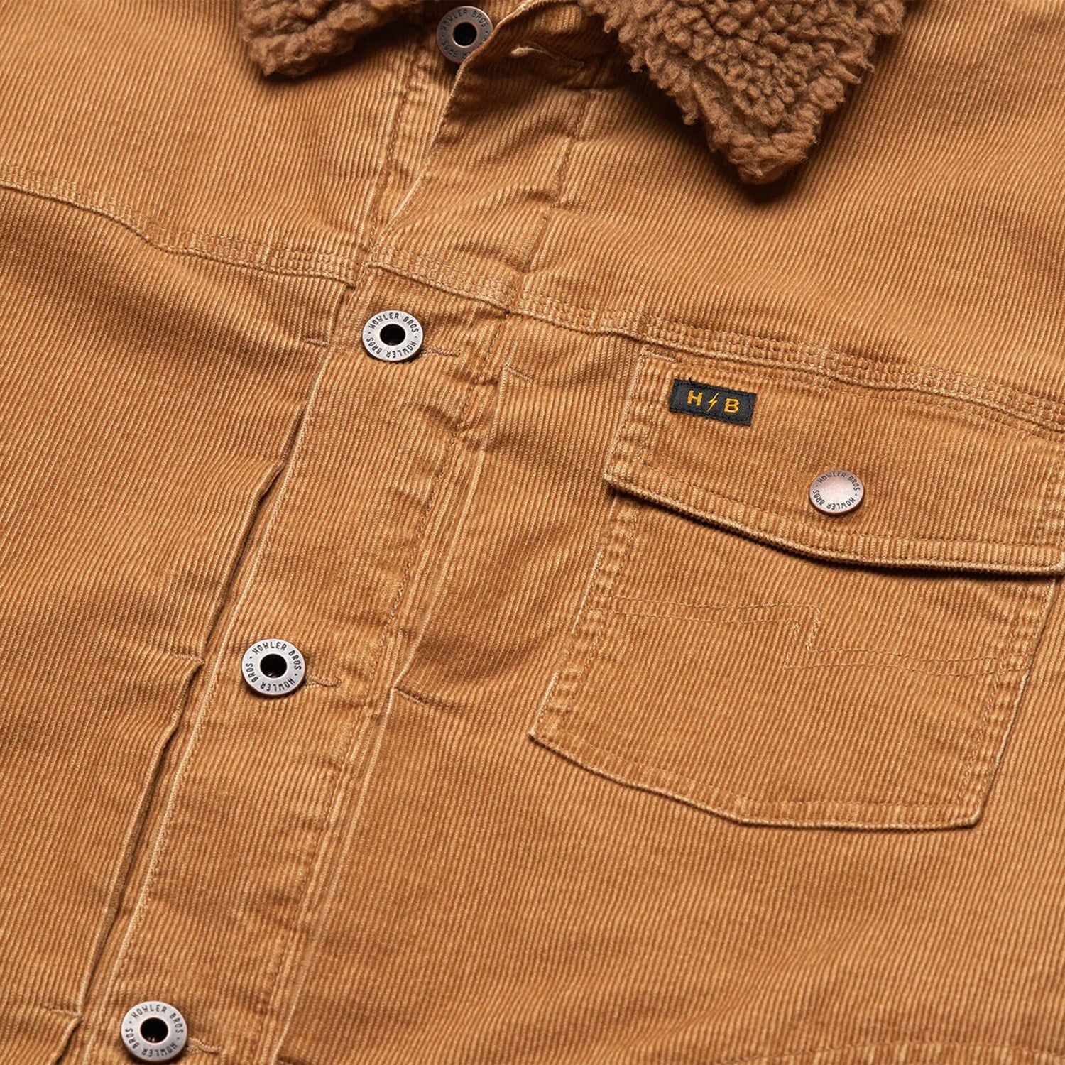 Close-up of a tan corduroy jacket, known as the Fuzzy Depot Jacket, featuring metal buttons, a brown sherpa collar, chest pocket, sherpa lining for warmth, and a small yellow “H.B” label above the pocket. - View 6