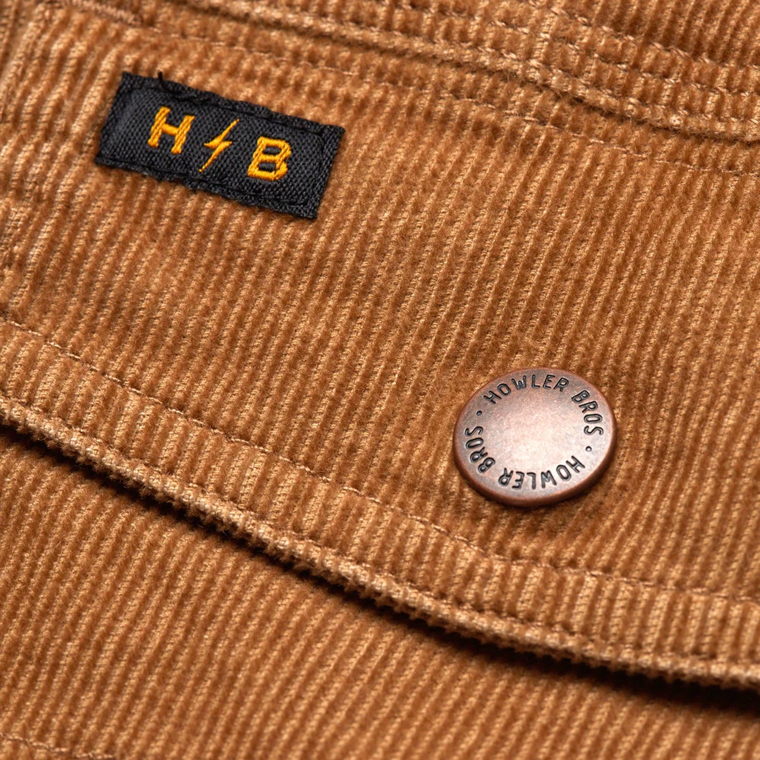 Close-up of brown corduroy jacket fabric with a black and yellow “H//B” label and a metal button engraved with “HOWLER BROS.”—a signature detail on the Fuzzy Depot Jacket. - View 8