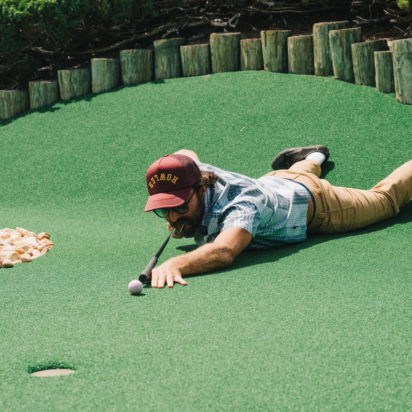 A man lies on his stomach on artificial turf at a mini-golf course, stretching out to putt a golf ball with his club held in his mouth. He wears a vintage-inspired hat, sunglasses, a plaid shirt, and khaki pants. - View 2