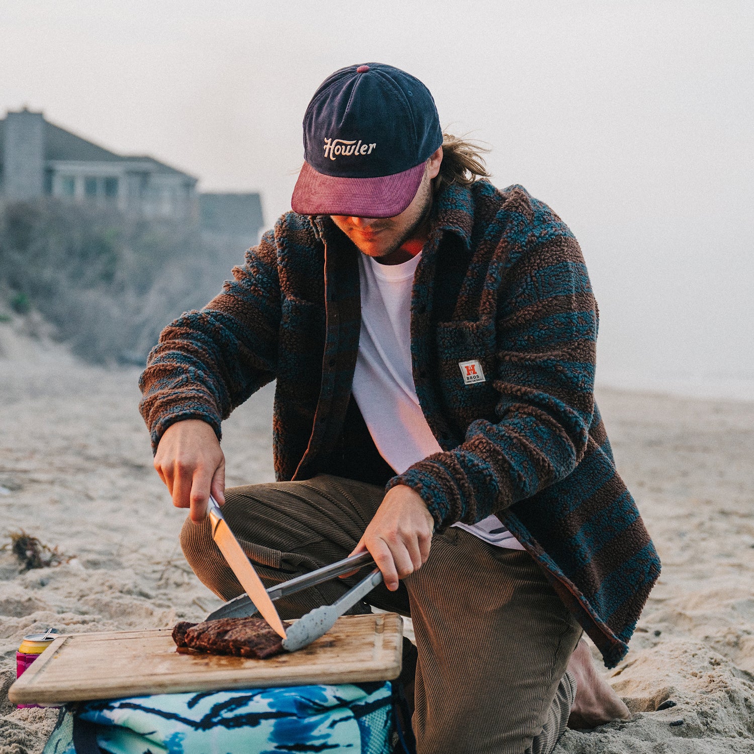 A person wearing an unstructured snapback with a soft crown and a striped jacket slices cooked meat on a wooden board at a sandy beach, with houses and the ocean in the background. - View 2